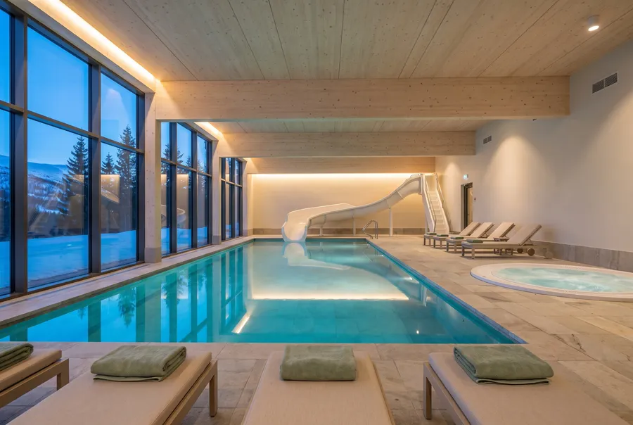 A heated indoor pool with a curving white waterslide and a round hot tub under a pale wood ceiling, loungers with sage towels, a window wall onto snowy spruce at dusk
